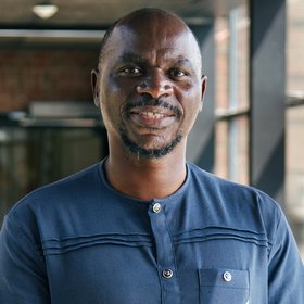 [Translate to English:] Porträt-Foto des gabonesischen Forschers Ghyslain Mombo-Ngoma: Er trägt Glatze, einen kurzen Vollbart und ein dunkelblaues afrikanisches Shirt und lächelt in die Kamera.