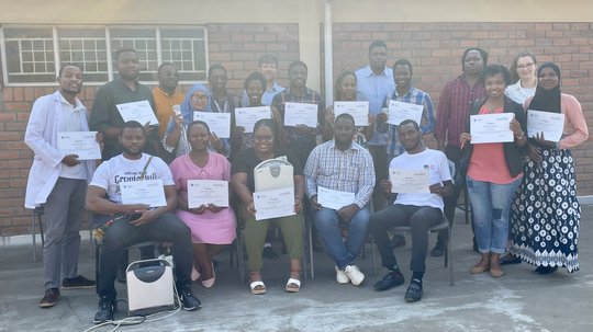 The picture shows a group of twenty people standing and sitting in front of a brick building. Some people are holding certificates and ultrasound machines.