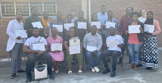 Das Bild zeigt eine Gruppe von zwanzig Personen, die vor einem Backsteingebäude stehen und sitzen. Einige Personen halten Urkunden und Ultraschallgeräte in der Hand.