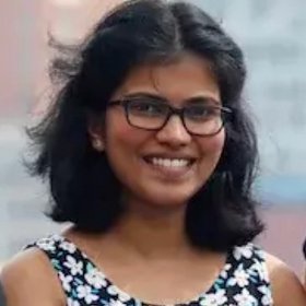 Shindhuja Joel A woman with dark, shoulder-long hair and wearing dark framed glasses smiling kindly into the camera.