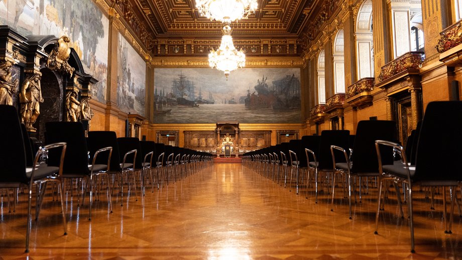 Foto vom Großen Festsaal des Hamburger Rathauses mit zwei Kronleuchtern an der Decke, edlem Parkett, reicher Bestuhlung links und rechts und historischen Wandgemälden.