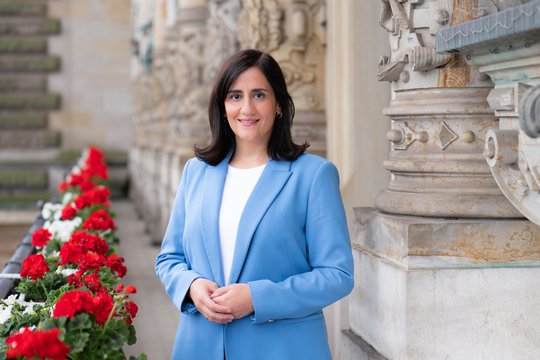 Maryam Blumenthal Hamburgs Wissenschaftssenatorin Maryam Blumenthal auf dem Balkon des Rathauses. Links rote und weiße Geranien, rechts eine Frau mit schulterlangen schwarzen Haaren, weißem Shirt unter hellblauem Blazer und freundlichem Blick.