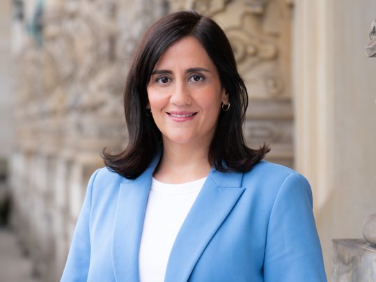 Maryam Blumenthal, Minister for Science, Research, and Equalities Hamburg Maryam Blumenthal, Minister for Science, Research, and Equalities Hamburg, On the balcony of the city hall. To the left, red and white geraniums; to the right, a woman with shoulder-length black hair, wearing a white shirt under a light blue blazer and a friendly expression.