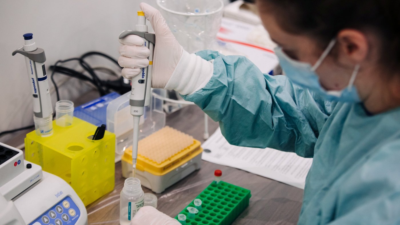 One scientist wearing gloves and a gown pipetting a liquid from a bottle to small tubes on a laboratory bench.