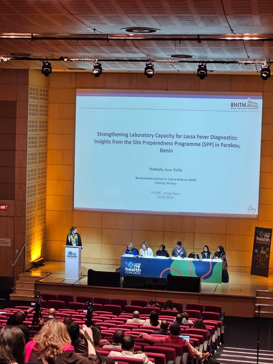 Picture from inside an auditorium with a diverse panel, a female speaker and the first slide of the BNITM presentation about SPP in Benin