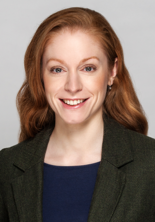 Ein Porträtfoto einer jungen Frau mit langen roten Haaren lächelnd freundlich. Sie trägt einen dunklen Blazer.