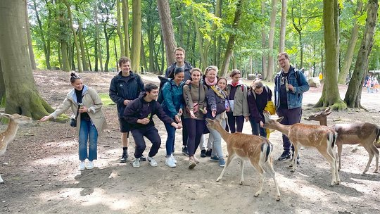 Auf dem Foto steht die Arbeitgruppe in einem Wald des Wildparks Schwarze Berge. Einige Mitglieder füttern vier Rehe, die im Vordergrund zu sehen sind.