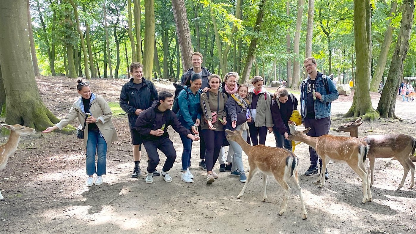 Auf dem Foto steht die Arbeitgruppe in einem Wald des Wildparks Schwarze Berge. Einige Mitglieder füttern vier Rehe, die im Vordergrund zu sehen sind.