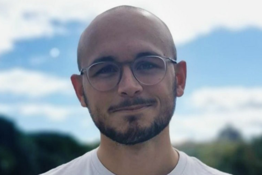 Louis Sarrazin: Ein Doktorand steht lacht freundlich in die Kamera. Er trägt eine Glatze, einen schwarzen Kinn- und Oberlippenbart, ein weißes Shirt und eine runde Brille. Er steht draußen vor einem blauen, aber leicht wolkigem Himmel.