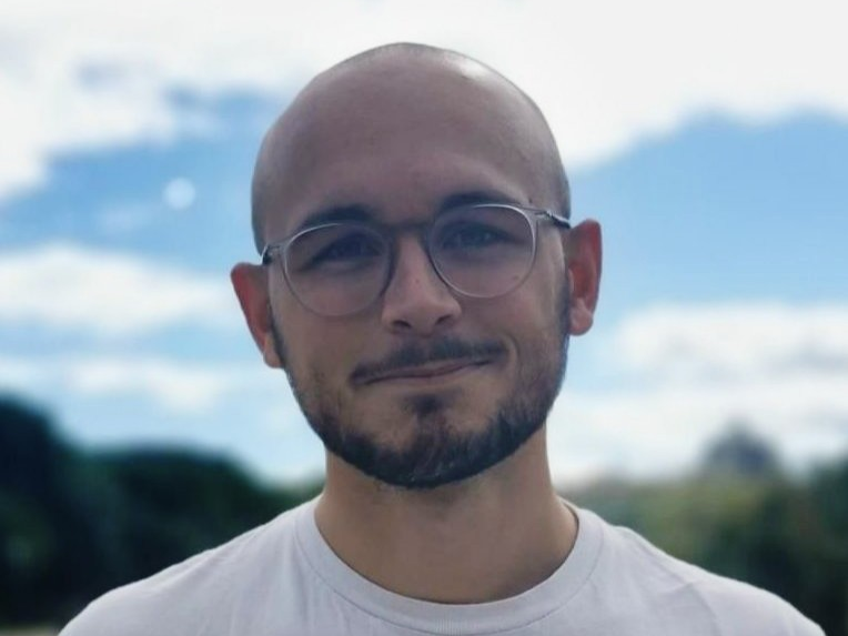 Louis Sarrazin Louis Sarrazin: Ein Doktorand steht lacht freundlich in die Kamera. Er trägt eine Glatze, einen schwarzen Kinn- und Oberlippenbart, ein weißes Shirt und eine runde Brille. Er steht draußen vor einem blauen, aber leicht wolkigem Himmel.