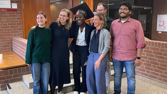 Jennifer Antwi-Ekwuruke's defense Eine gemischte Gruppe von sechs Personen steht lächelnd und dicht beieinander in einem Raum vor einer Backsteinmauer. Sie wirken fröhlich und lässig. Einer von ihnen trägt einen Doktorhut.