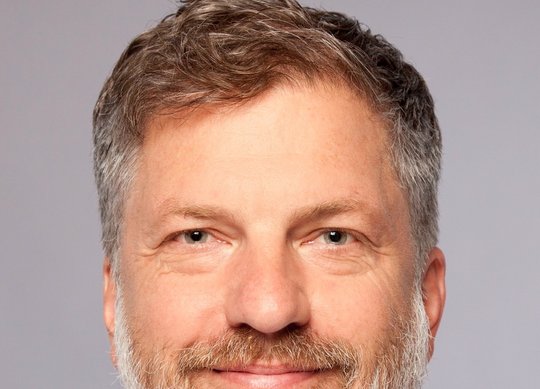 Dr. Robin Kobbe: ein Arzt mit kurzen grauen Haaren, trägt ein graues Hemd und steht vor einer dunklen Backsteinmauer.