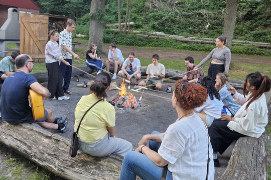 LCI Summer School 2026 Grillen Das Foto zeigt ein paar Studierende draußen beim Grillen. Einige sitzen auf Baumstämmen.