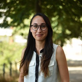 Dr. Emily Nelson Dr. Emily Nelson: eine lächelnde Forscherin mit langen dunklen Haaren und Brille. Sie trägt eine weiße Bluse mit schwarzer Knopfleiste und steht auf einem sonnigen Weg mit Bäumen.