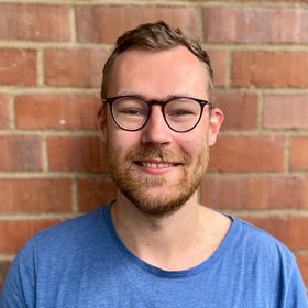 Lennart Sänger: A PhD student smiles at the camera. He has short dark blond hair, round horn-rimmed glasses, a short beard and a blue shirt.