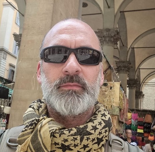 Bernhard Zibrat Bernhard Zibrat: Ein technischer Assistent schaut in die Kamera. Er trägt eine Sonnenbrille, einen Glatze, einen grauen Bart und einen hellgelben Schal mit Sternmuster. Im Hintergrund ist ein antiker, aus Sandstein gebaueter Durchgang mit Deckenbögen zu sehen.