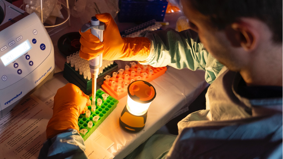The image shows a lab bench lit by a camping light while a staff member pipettes samples into tubes in a green rack.