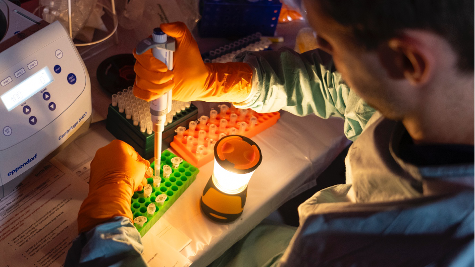 EMLab member preparing samples in the field laboratory The image shows a lab bench lit by a camping light while a staff member pipettes samples into tubes in a green rack.