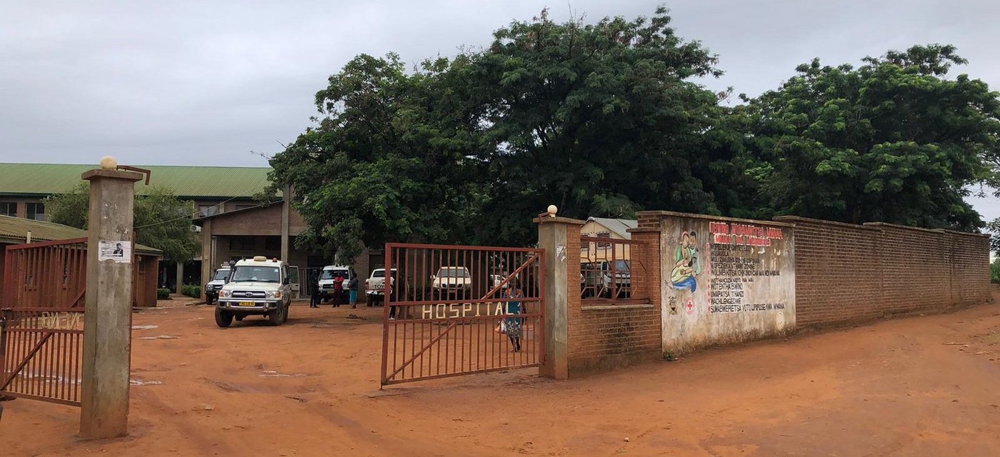 Das Bild zeigt das Distriktkrankenhaus in Neno, Malawi. Zu sehen ist das offene Tor zum Eingang und die Mauer drumherum. Der Boden ist lehmfarbig und es stehen ein paar Autos und Menschen vor dem Krankenhaus.
