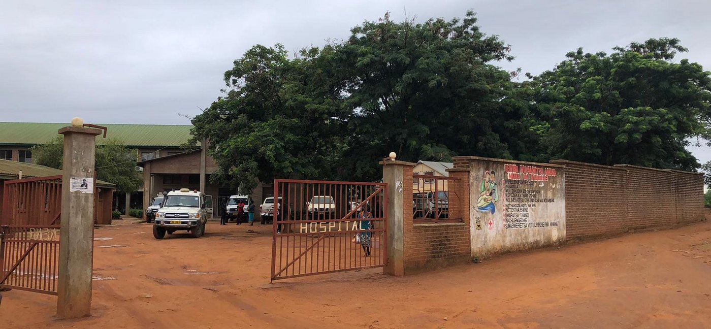 The picture shows the district hospital in Neno, Malawi. The open gate to the entrance and the wall around it can be seen. The ground is clay-coloured and there are a few cars and people standing in front of the hospital.