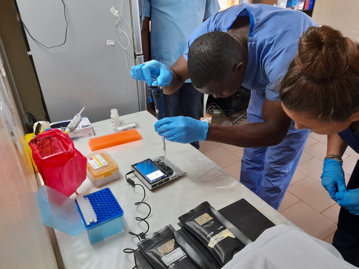 One lab staff from Gueckédou lab loading sequencing samples in a MinION flow cell (Oxford Nanopore Technologies) during a joined metagenomics sequencing training at CRV-LFHVG. May 2024, Conakry, Guinea A lab staff is pipetting a sample into a sequencing device, watched by a colleague. He is wearing a blue lab suit and bright blue gloves, red, orange, yellow and blue lab equipment is on the table.