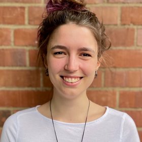 Saskia Johanns: A doctoral student laughs into the camera. She wears her brown hair tied in a messy bun and a white shirt.