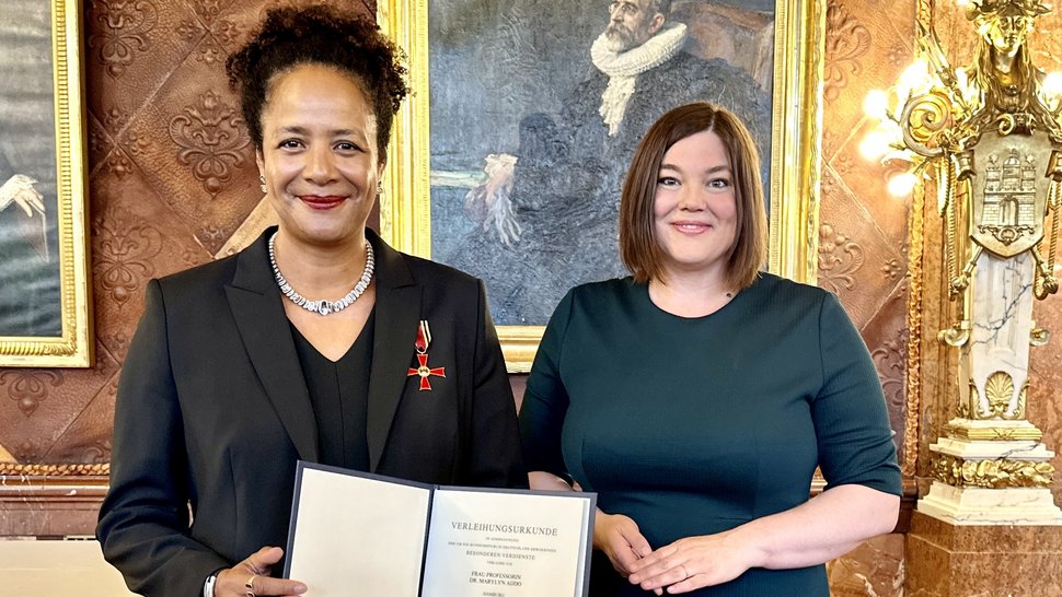 Das Foto zeigt links Frau Prof. Marylyn Addo mit der Urkunde in der Hand und dem Bundesverdienstorden an der Brust, rechts Wissenschaftsenatorin Katharina Fegebank. Beide lächeln. Sie stehen vor einer festlichen historischen Wand im Rathaus.