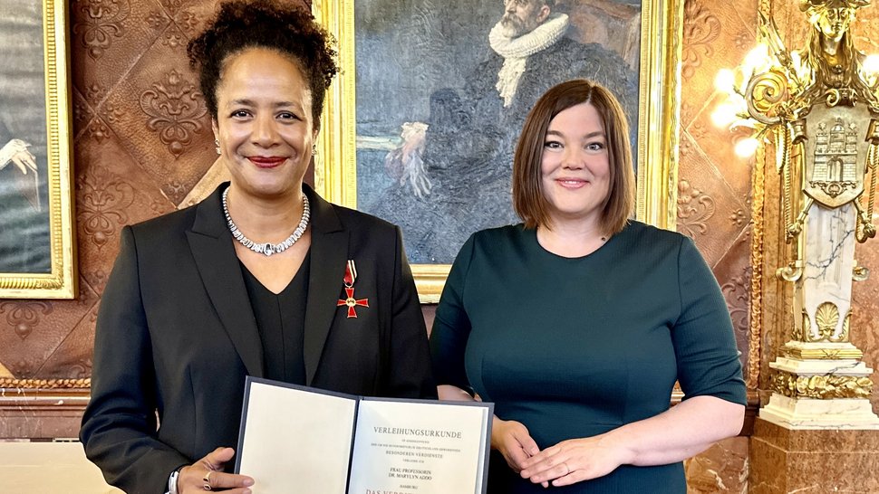 The photo shows Prof. Marylyn Addo on the left with the certificate in her hand and the Order of Merit on her chest, and Science Senator Katharina Fegebank on the right. Both are smiling. They are standing in front of a festive historical wall in the City Hall.