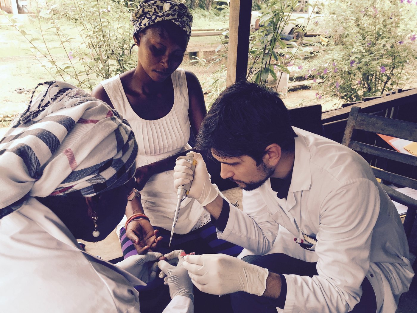 A doctor examines a person on a covered, but still open terrace while another observes him.