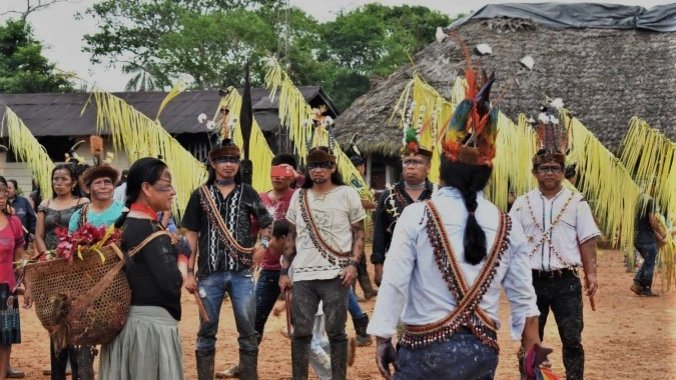 Quechua indigenous people in Sarayaku near Río Bobonaza/ Ecuador. Eine Gruppe Menschen Kopf- und Körperschmuck, sowie Gesichtsbemalung stehen auf einem Platz. Eine Frau trägt einen Korb mit bunten Blättern und Blumen auf dem Rücken und einige Menschen tragen große gelbe Palmenwedel in den Händen.
