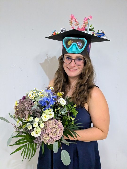 the picture shows charlotte hansen with her doctoral hat and a bouquet of flowers after completing her dissertation. A pair of diving goggles is attached to the hat, which was made by her colleagues, and on top you can see aquarium plants, a syringe and antibody tubes
