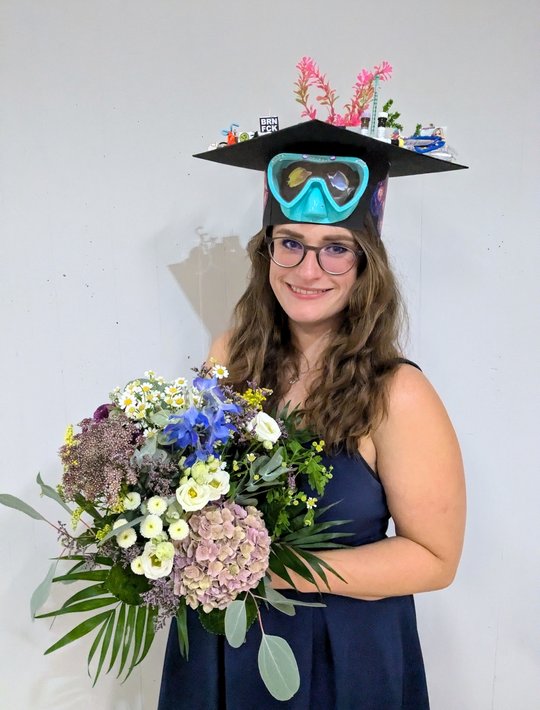 the picture shows charlotte hansen with her doctoral hat and a bouquet of flowers after completing her dissertation. A pair of diving goggles is attached to the hat, which was made by her colleagues, and on top you can see aquarium plants, a syringe and antibody tubes