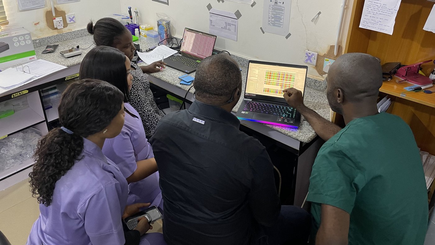 Sequencing lab staff performing bioinformatics analysis of sequencing results to obtain sequences alignments for phylogeny. February 2024, Irrua, Nigeria. The photo shows five Nigerian colleagues at a lab bench with two laptops on it. They are looking at one of the screens on which a bioinformatic analysis is being performed.
