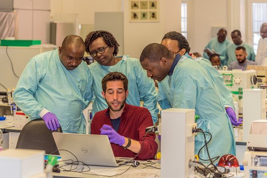 Group of researchers in a lab wearing lab uniforms surrounding another researcher and his laptop as he explains something to them.