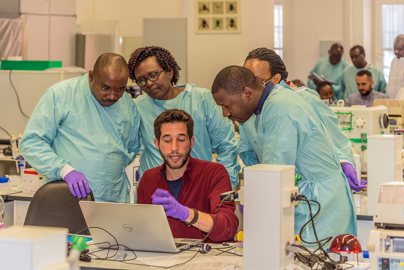 Gruppe von Forschern in einem Labor in Laborkleidung, die einen weiteren Forscher und seine Laptop umringen,während dieser ihnen etwas erklärt.