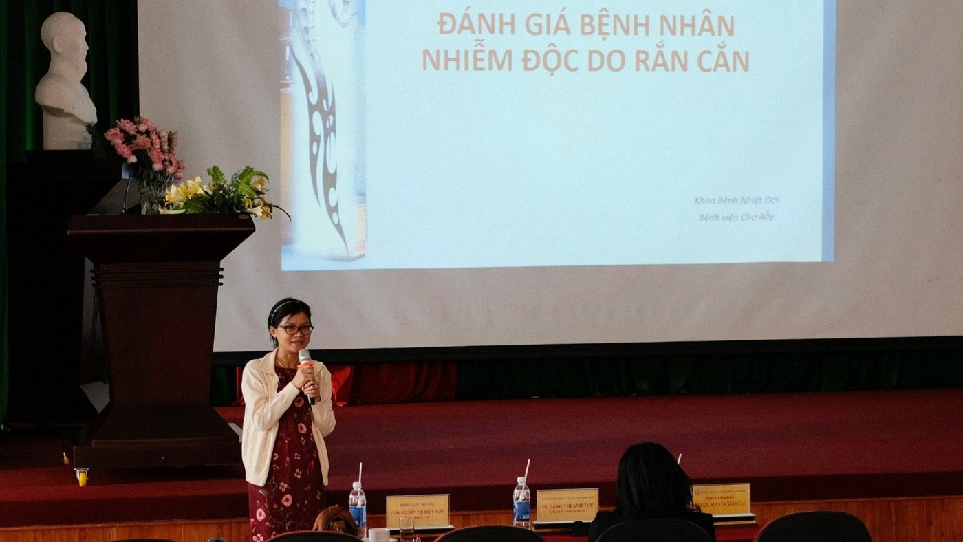 Das Bild zeigt eine Frau, die in ein Mikrofon spricht und vor einer projizierten Präsentation steht. Auf der Präsentation ist vietnamesische Schrift zu sehen.