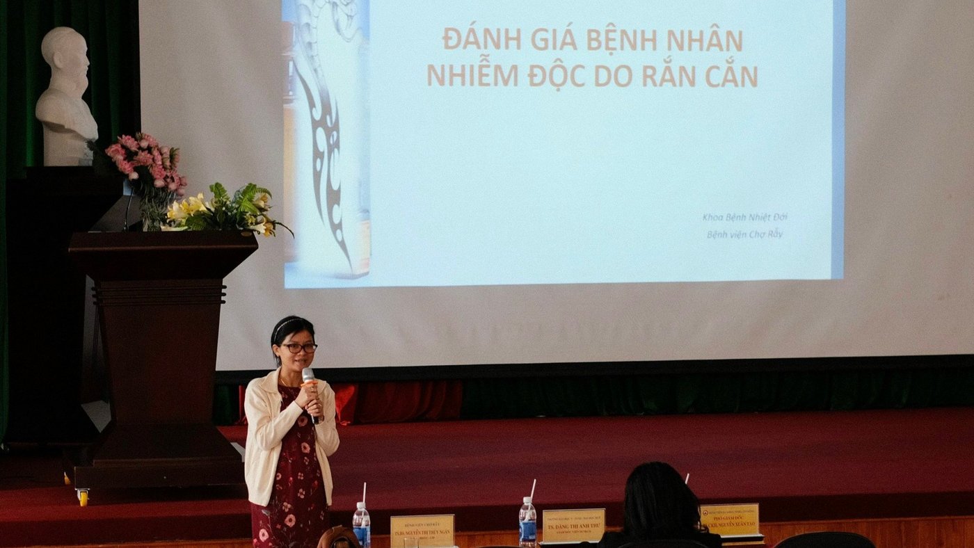 The picture shows a woman who is speaking into a microphone and standing in front of a projected presentation. On the presentation is Vietnamese writing.