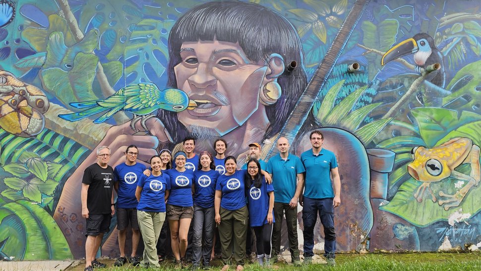 Das Foto zeigt eine Gruppe Menschen vor einem überlebensgroßen bunten Wandgemälde: Ein indigenes Gesicht aus Peru, umgeben von Urwald, in dessen Mund gerade ein Papagei fliegt. Passend zum One-Health-Gedanken.