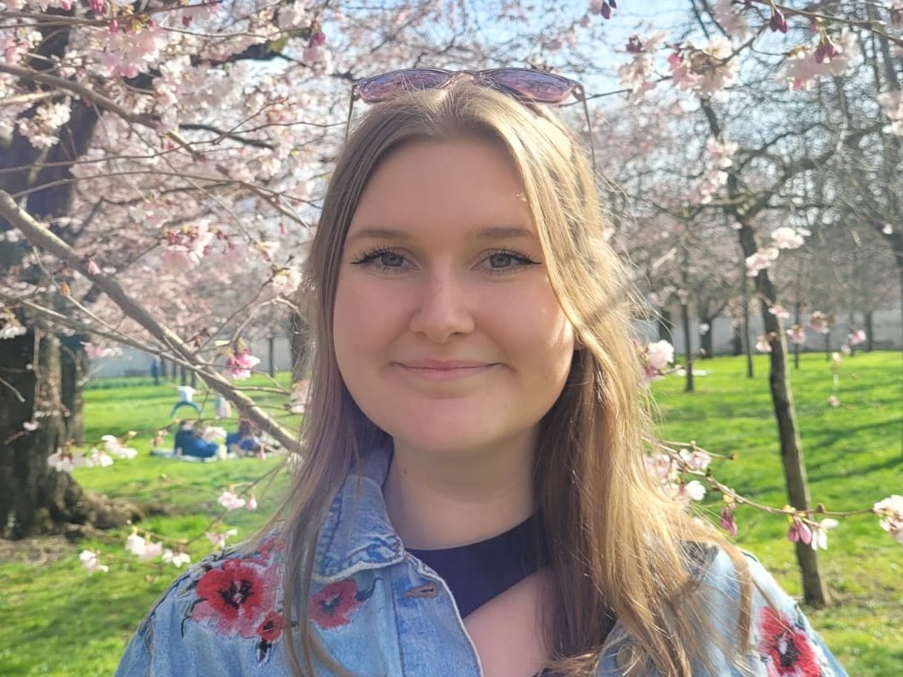 Fenja Nuglisch Eine junge Frau steht vor einer Wiese mit blühenden Bäumen und lächelt freundlich in die Kamera.