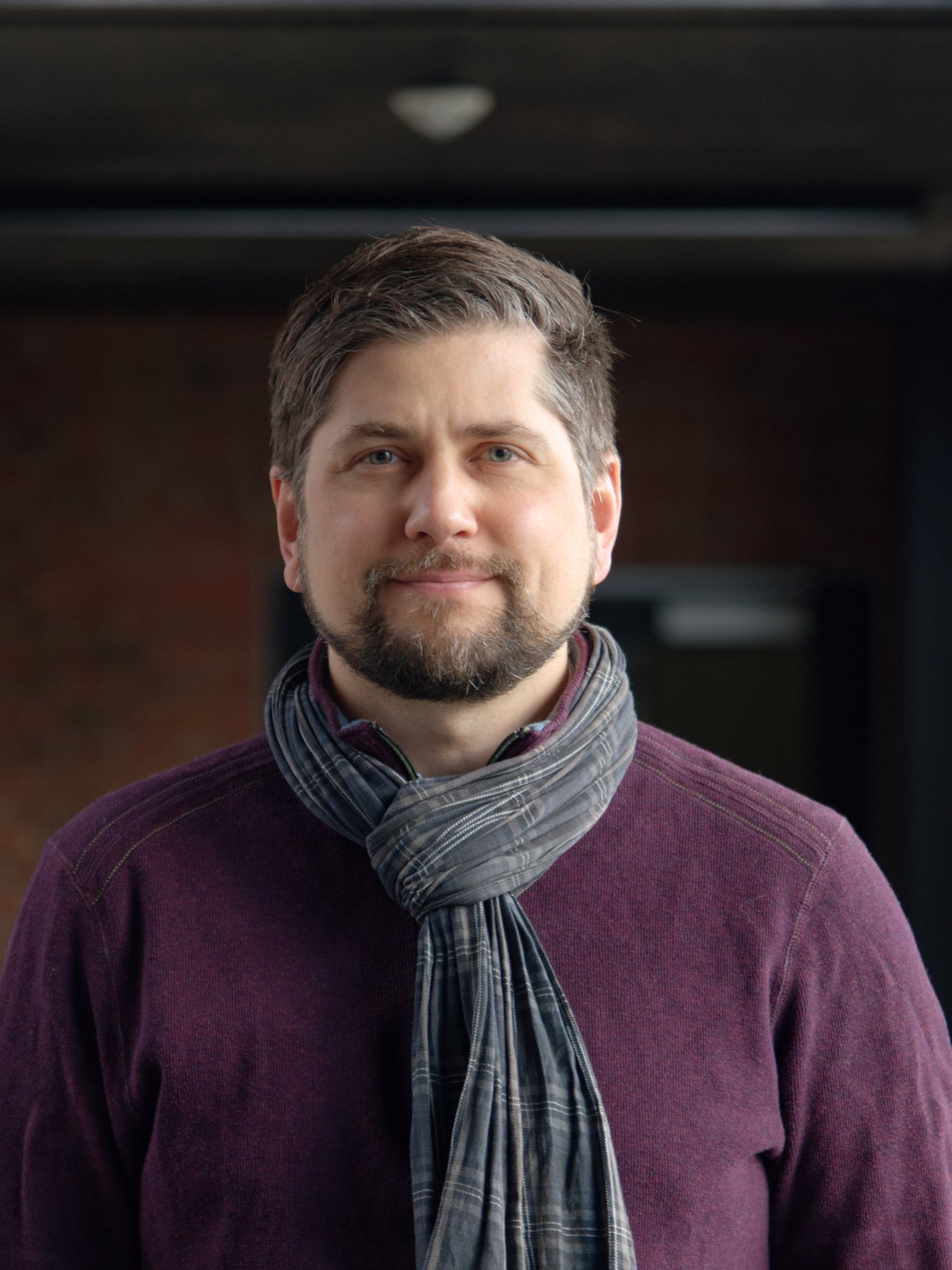 An experienced researcher can be seen smiling friendly into the camera. He wears short brown-grey hair gelled back, a light beard, a scarf and a purple colored top.