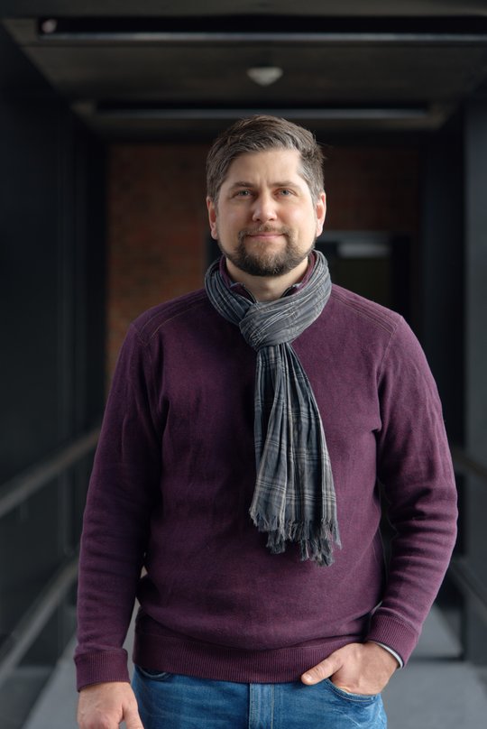 Dr. Johannes Mischlinger: ein Gruppenleiter, der kurzes, nach hinten gegeltes braun-graues Haar,  einen leichten Bart, einen Schal und ein lila farbendes Oberteil trägt.