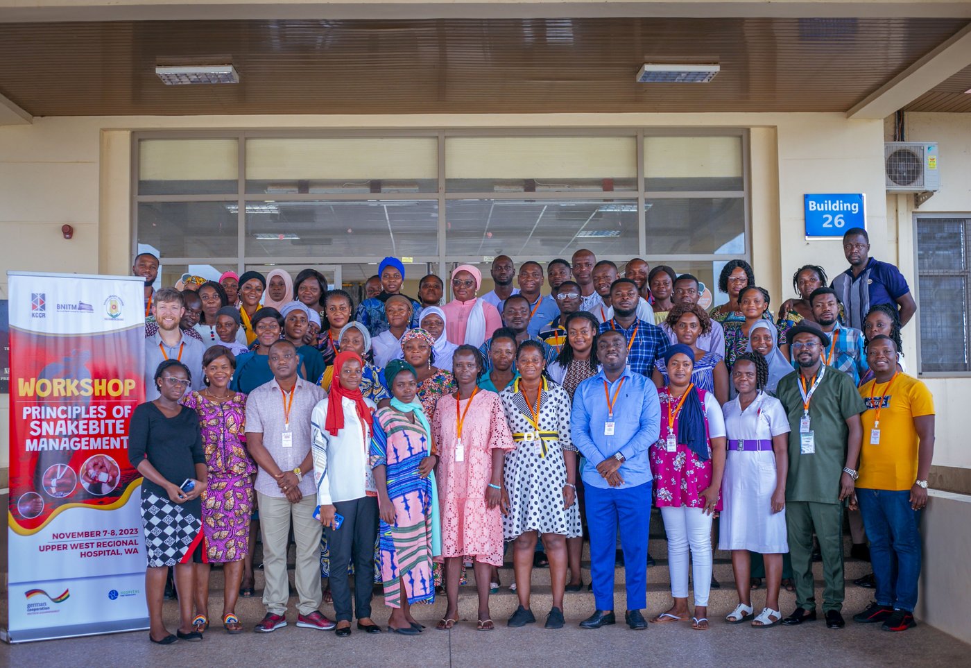 The picture shows a group of people standing in front of a hospital entrance next to a banner saying 'Workshop - Principles of Snakebite Management'. Everyone is looking friendly into the camera.