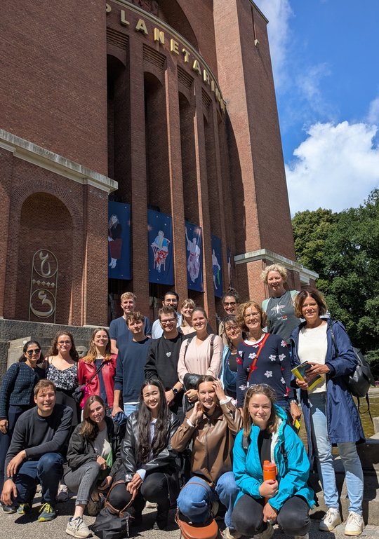 In the foreground, a group of 19 people are kneeling and smiling at the camera. In the background, you can see the front of a red brick building with large golden letters spelling out ‘Planetarium.’ Below that are colourful constellations displayed on posters.