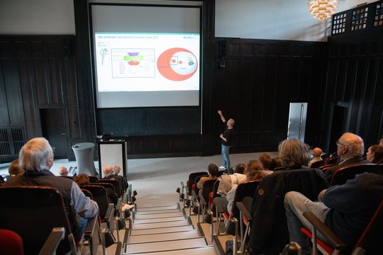 Auf dem Foto ist links und rechts das Publikum im Hörsaal des BNITM zu sehen. Mittig sieht man die Leinwand mit einer Folie des Vortrags. Vorne sieht man Tobias Spielmann, der einen Vortrag hält.