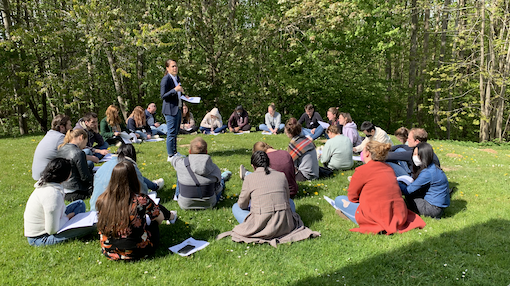 A group of young people sits in a circle on a meadow and exchanges ideas. In the middle is a woman in a suit with a piece of paper in her hand. The lecturer.