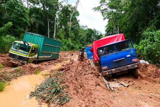 The photo shows two broken-down lorries on muddy roads in the rainforest. A person on a motorbike is riding close to one of the lorries.