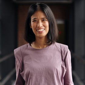 Dr. Eleonora Schönherr: a woman with shoulder-length dark hair and a lilac-coloured top stands in a glass passageway.