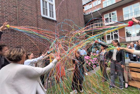 Barbara Honecker, who has just been awarded an honour, can be seen here in the garden with her colleagues, who shower her with streamers. She laughs, holds a drink in her hand, and bows her head.