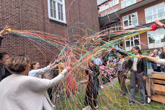 Barbara Honecker, who has just been awarded an honour, can be seen here in the garden with her colleagues, who shower her with streamers. She laughs, holds a drink in her hand, and bows her head.