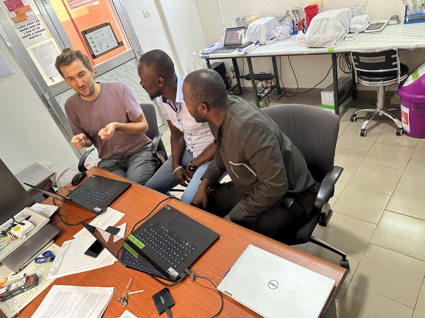 Bioinformatic training by BNITM at CRV-LFHVG. Two laboratory staff discussing with one trainer in front of two laptops used for bioinformatic analysis in the sequencing laboratory.